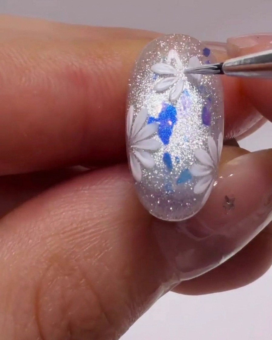 A fine liner brush creating precise white petal shapes on top of a shimmering, glittery nail.