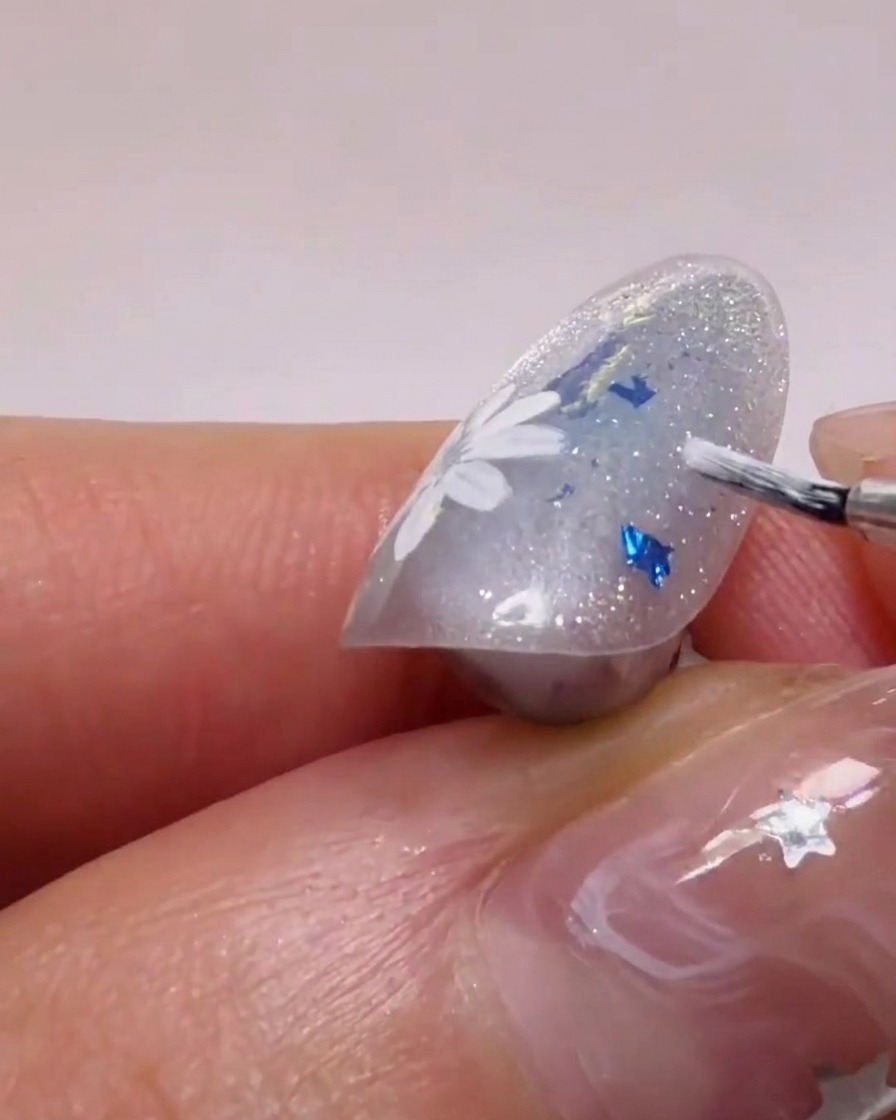 A close-up view of a nail brush applying white gel to form daisy petals over a glittery base.