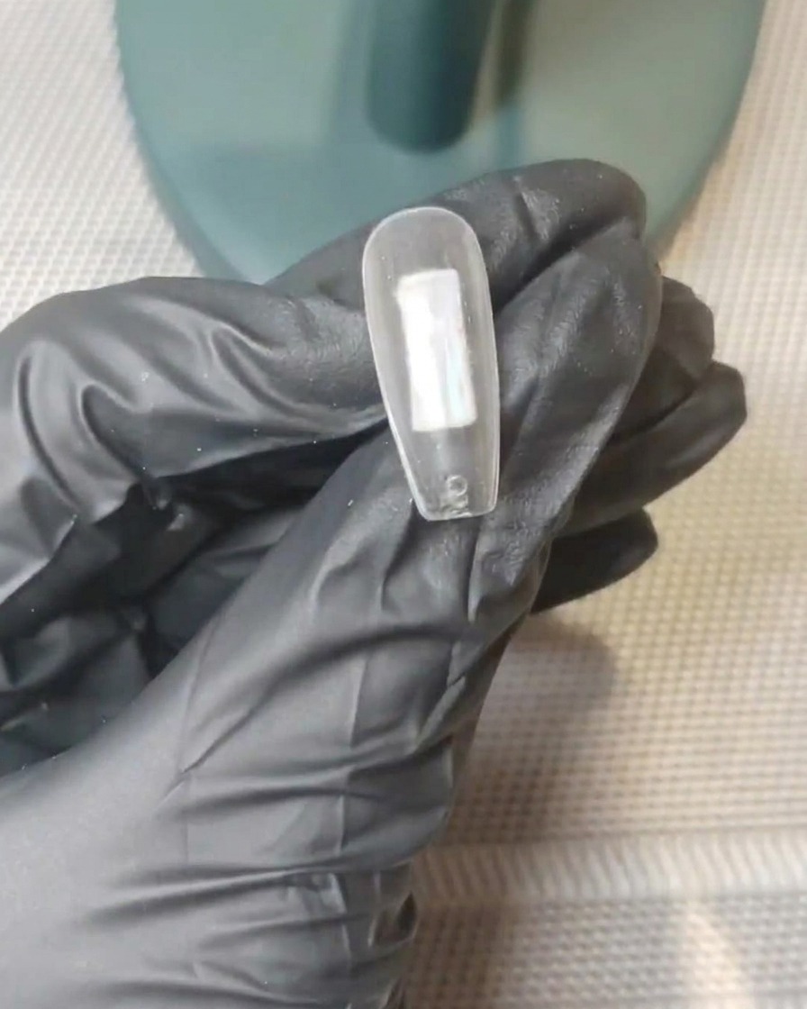 A close-up of a clear nail tip with distinct dots of nude pink and milky white gel placed on the center of the surface.