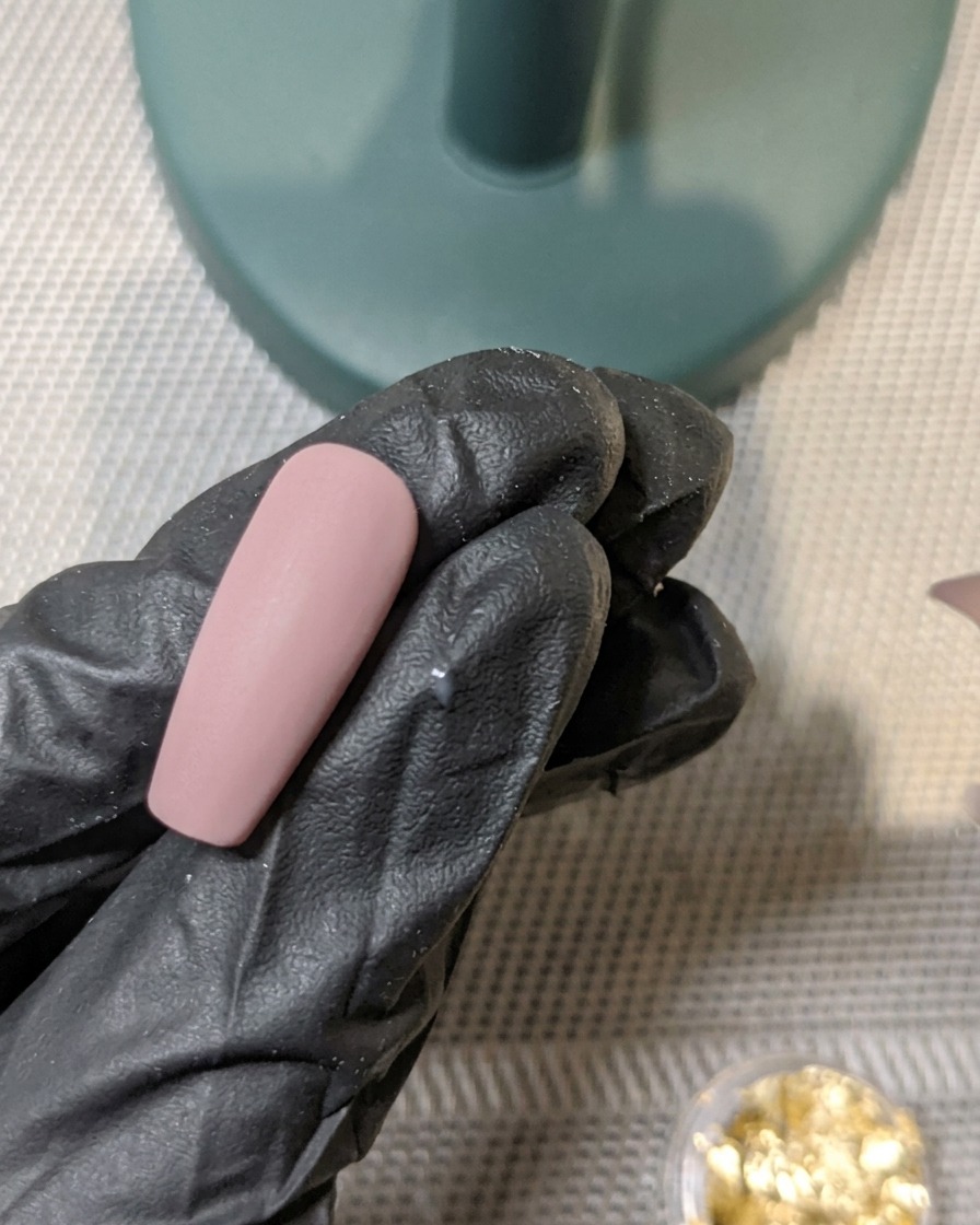 A brush applying a layer of matte top coat over a nail decorated with embedded gold foil.