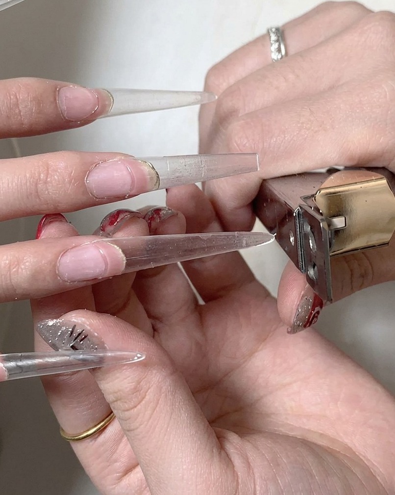 A nail brush applying a clear gel base coat across the transition point where the natural nail meets the extension.