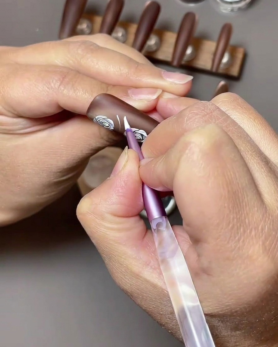 A close-up view showing white nail art gel being used to paint leaf shapes next to existing white roses on a brown nail.