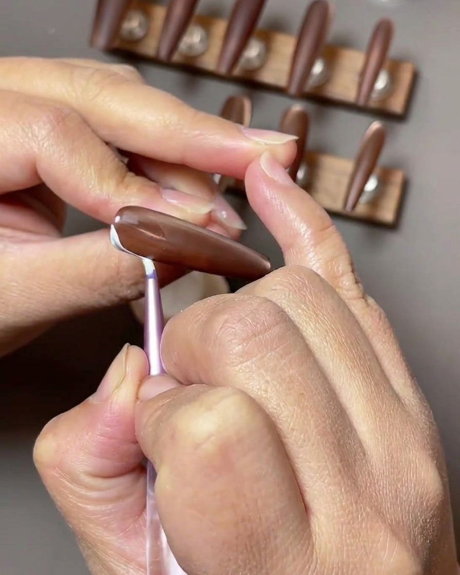Close-up of a brown nail where delicate white ribbon-like strokes are being painted along the side of the nail surface.