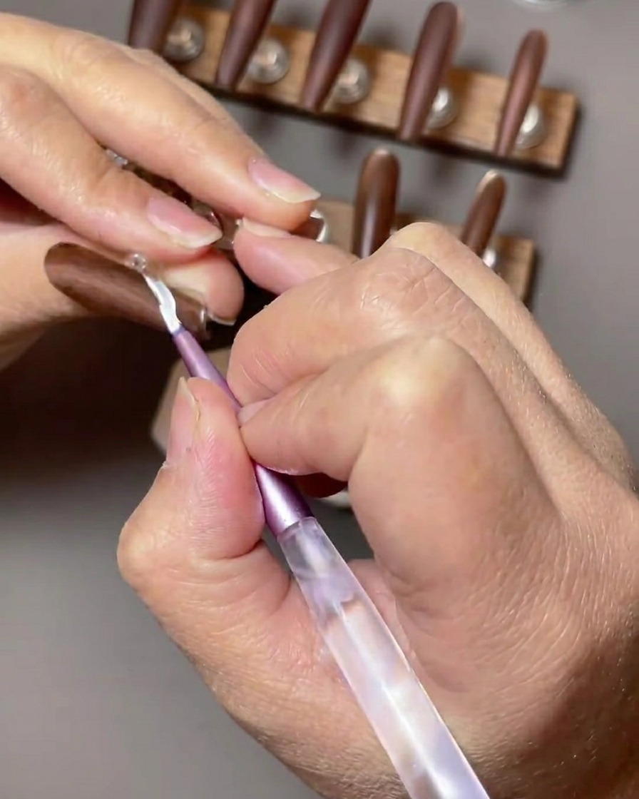 A close-up of a brown nail being accented with a white French tip design applied with a thin nail art brush.