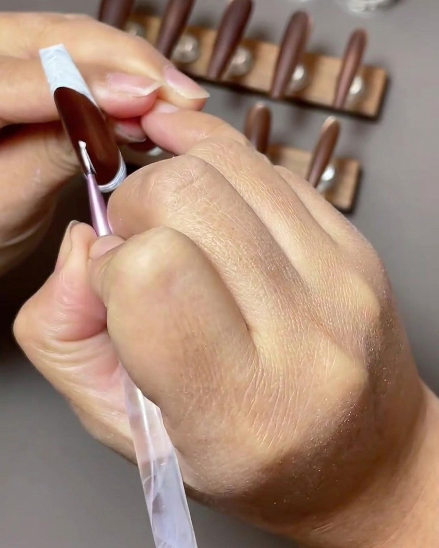 A close-up view of a thin nail art brush applying a white gel four-pointed star to a dark brown nail with a reverse French tip.