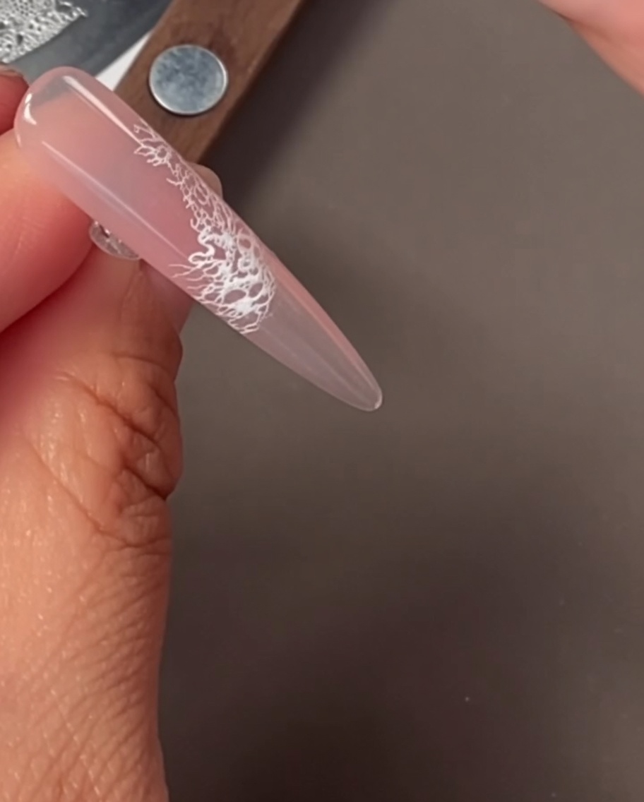 A close-up of a nail with a delicate white lace pattern applied via stamping.
