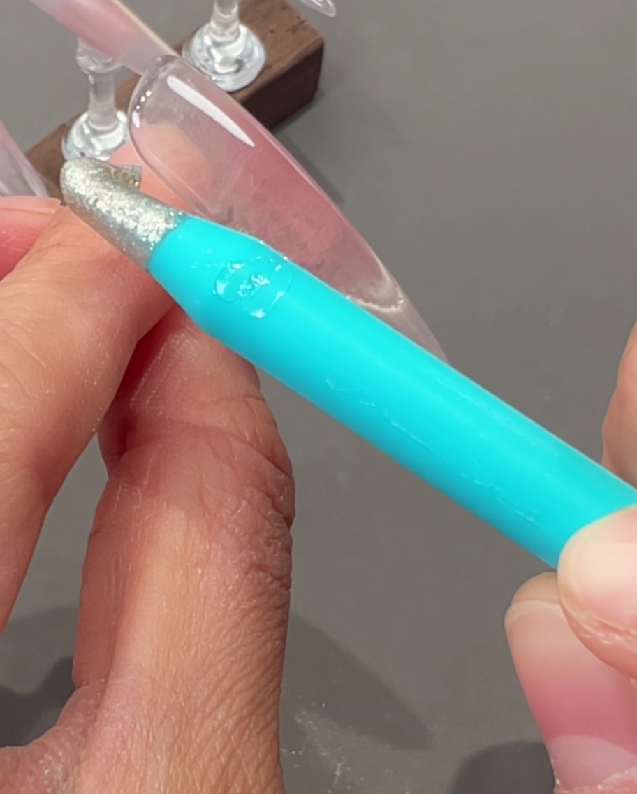 A hand holding a thin liner brush, drawing fine silver lines onto a nail surface.