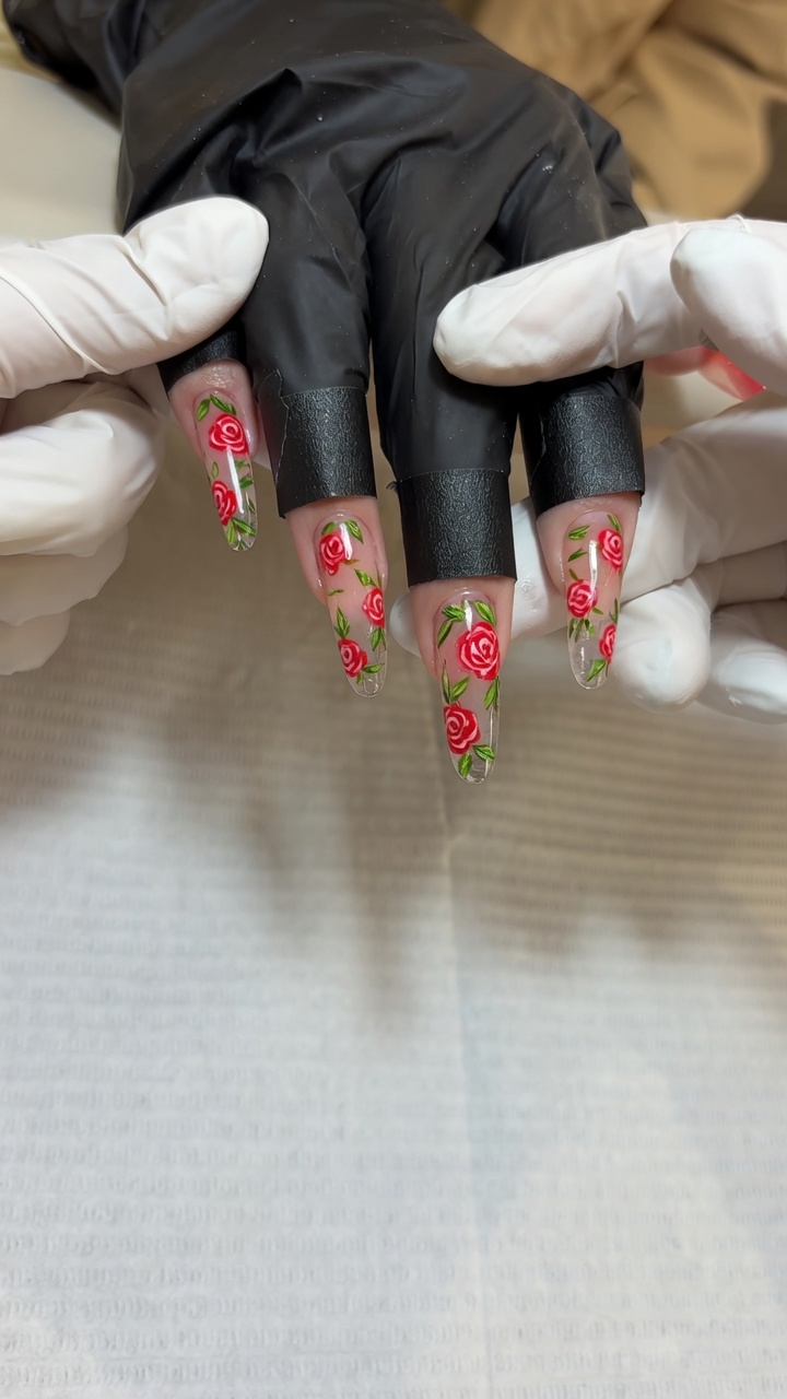 Close-up of long, clear gel nails featuring intricate, hand-painted red rose art and delicate green leaves.