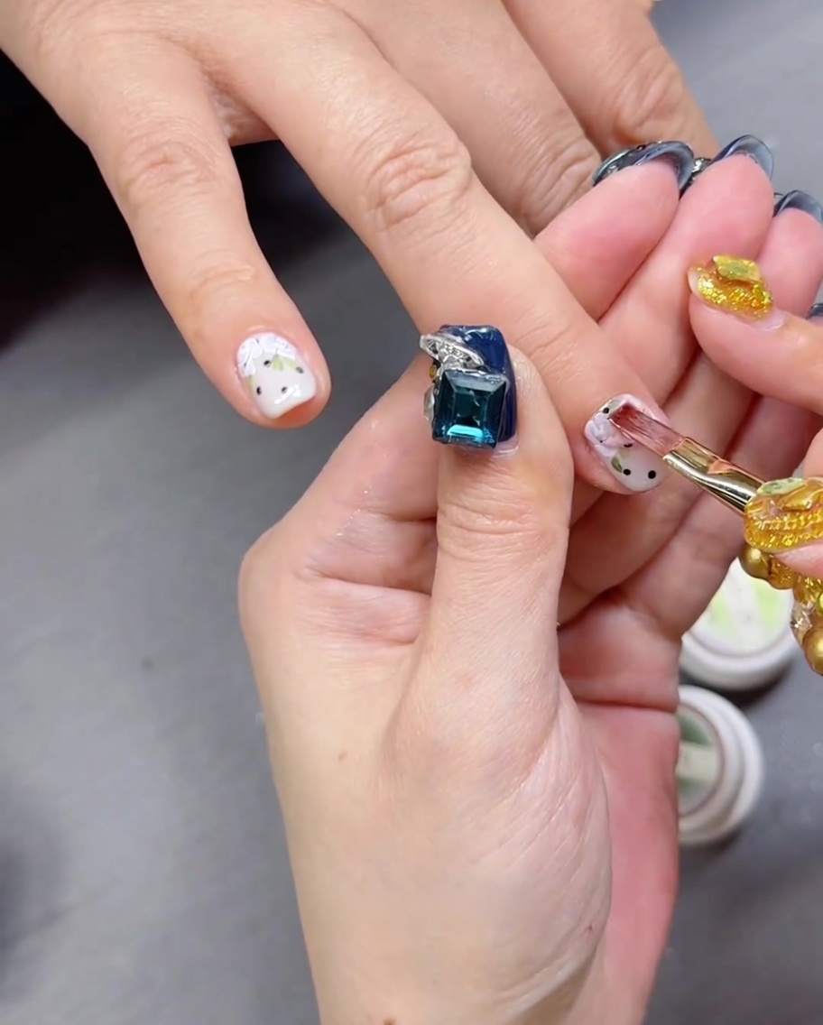 A close-up view of a brush applying a clear, glossy top coat over a nail featuring a hand-painted rose and polka dot design.
