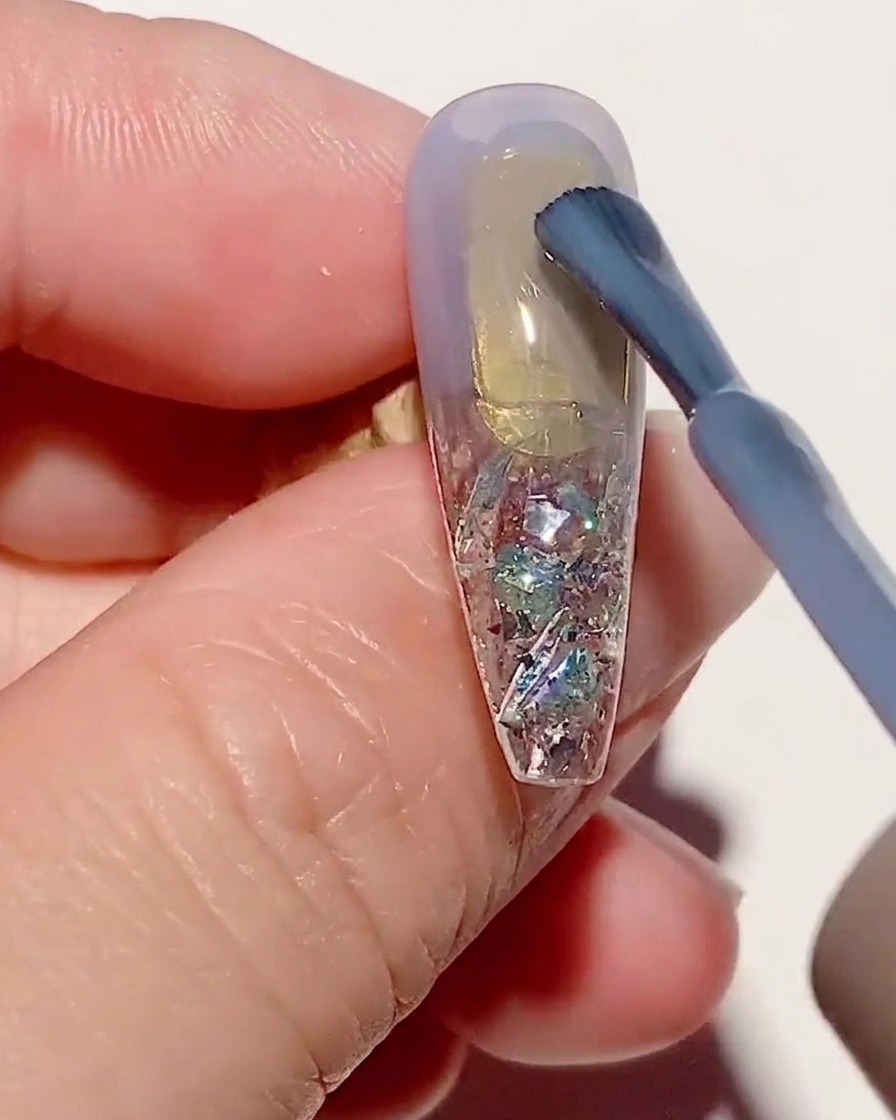 A brush applying a thin layer of sheer blue gel polish onto a textured nail tip.