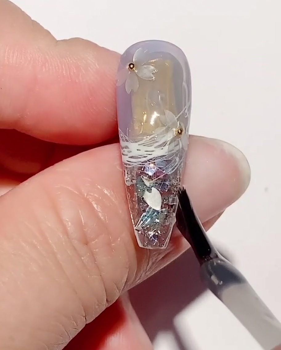 A nail art brush applying a layer of clear reinforcement gel over cherry blossom sequins and steel beads on a nail.
