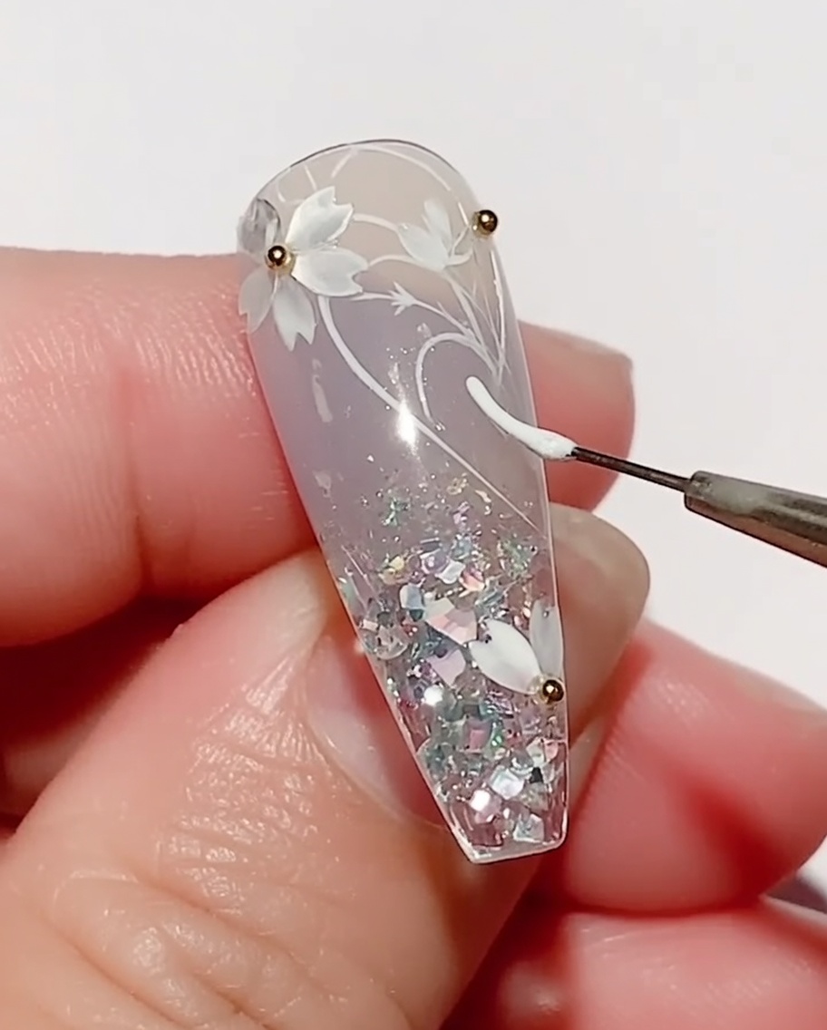 A thin liner brush applying white color gel to draw a leaf shape on a nail decorated with cherry blossoms.