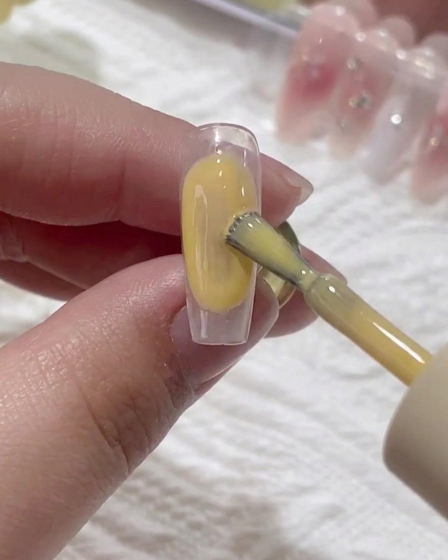 A brush applying a thin layer of clear base gel onto a natural fingernail.