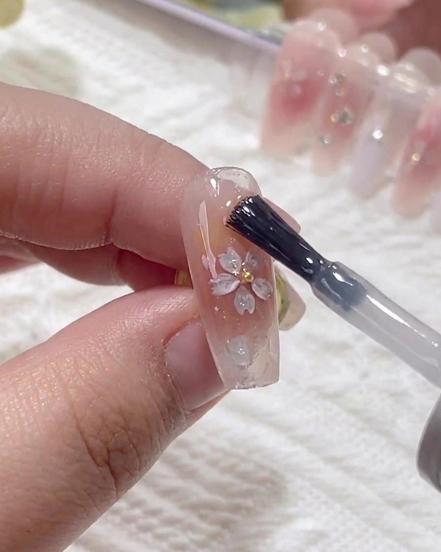 A brush applying a layer of clear strengthening gel over the nail surface, covering the applied cherry blossom sequins.