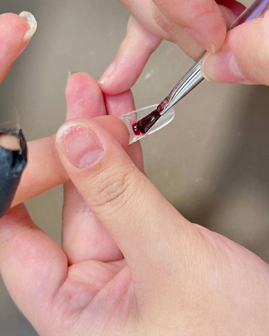 A brush applying a coat of sheer, milky white gel polish onto the surface of a clear nail tip.