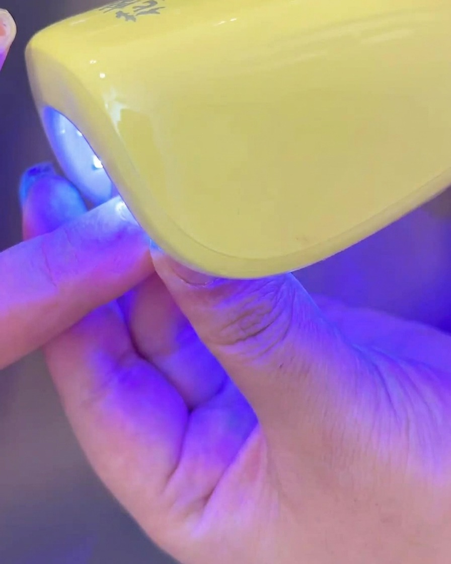 A hand placing a nail with applied milky white gel under a small UV/LED lamp for curing.