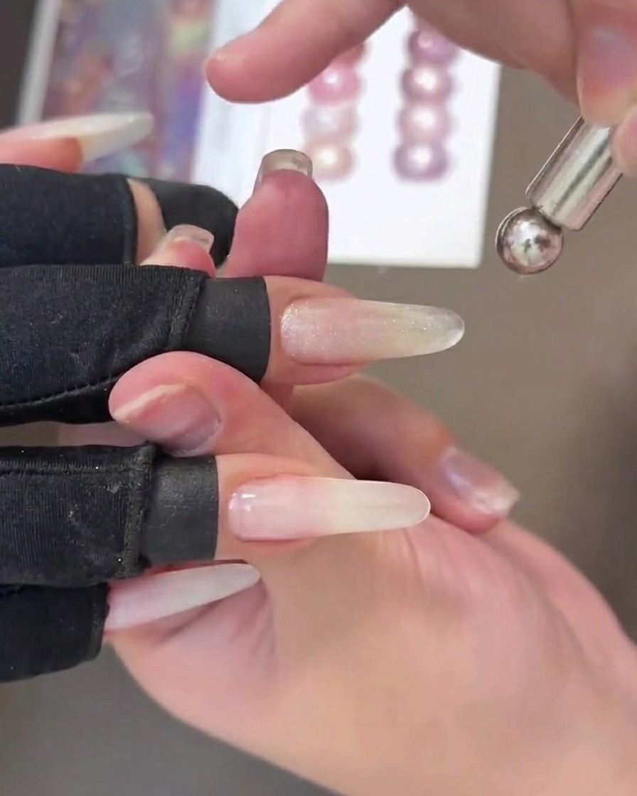 A close-up view of a nail with wet cat-eye gel, showing a metallic streak being pulled across the center by a handheld magnetic tool.