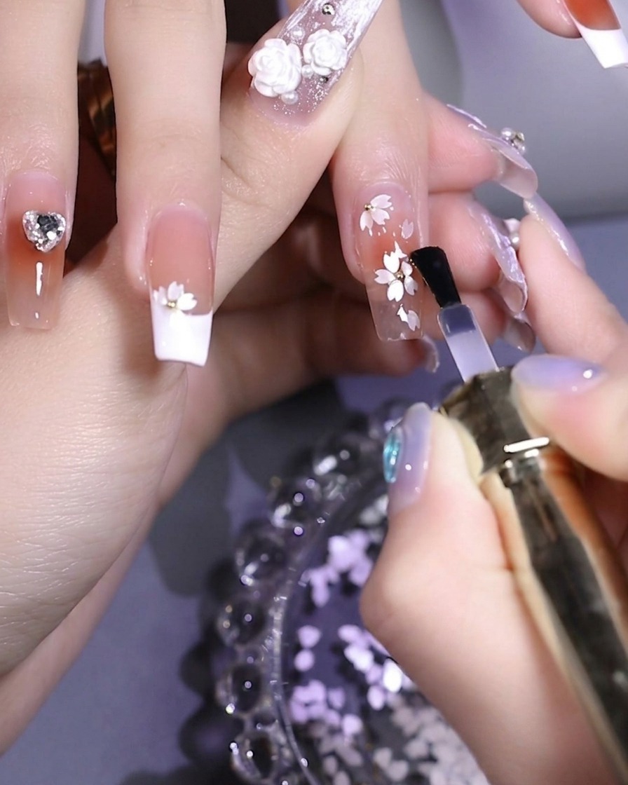 A brush applying a clear, glossy top coat over a nail decorated with white floral sequins and a central metal bead.