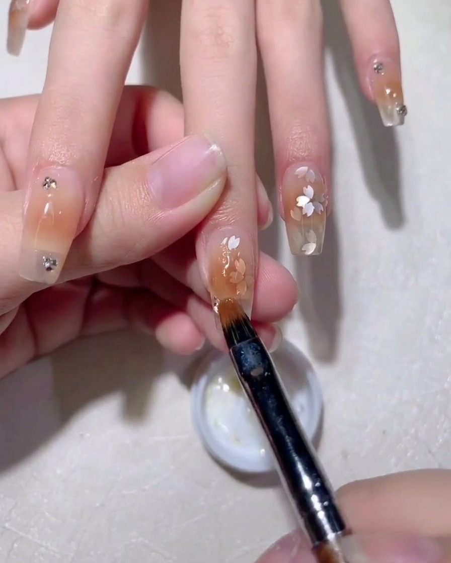 A close-up view of a nail art brush smoothing clear builder gel over delicate cherry blossom-shaped sequins on a nude-toned nail.