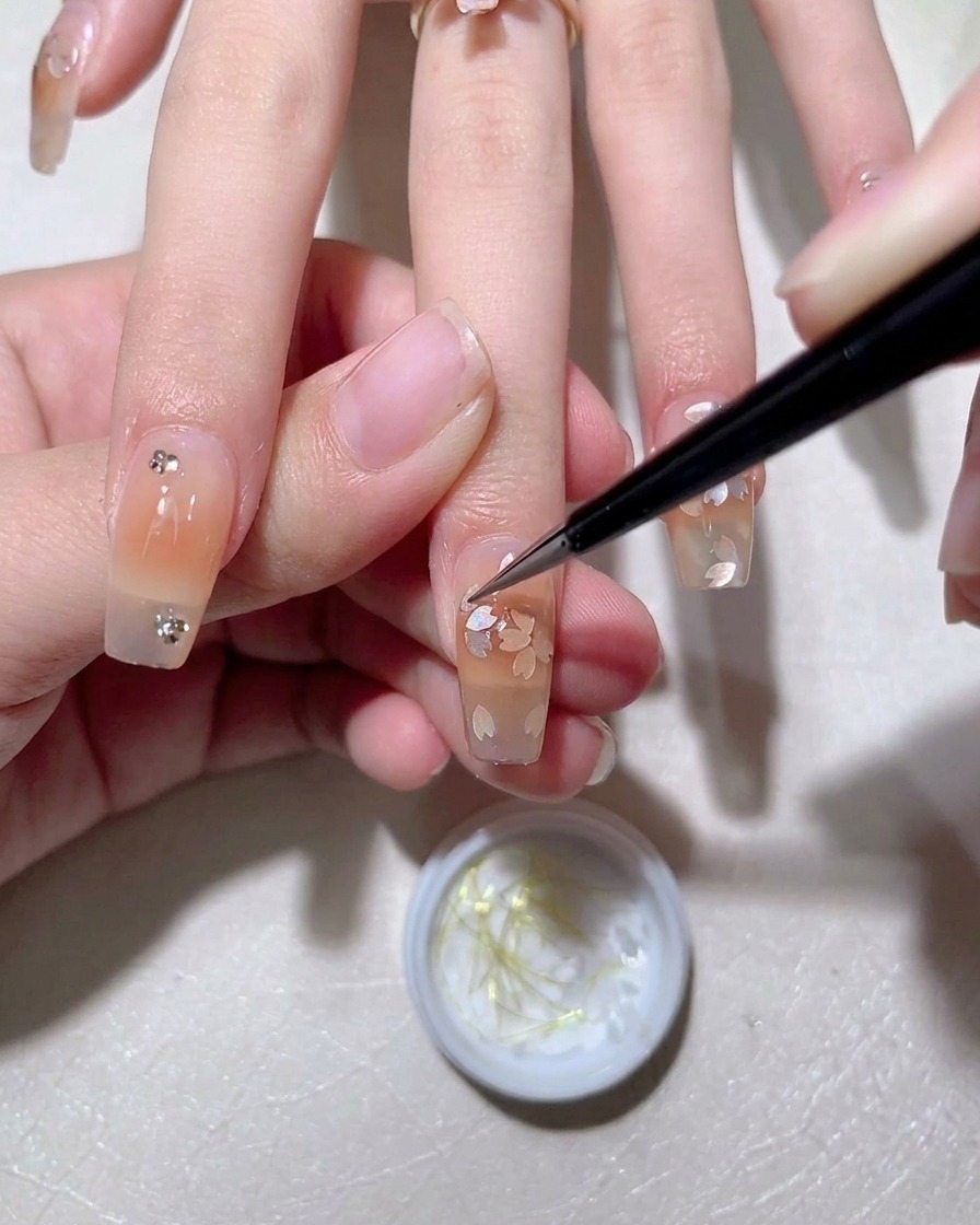 A detail brush positioning a small silver rhinestone on the nail next to a flower design.