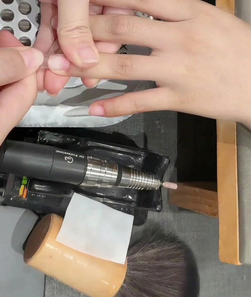 A nail buffer being used on the surface of a clear nail tip to remove shine.