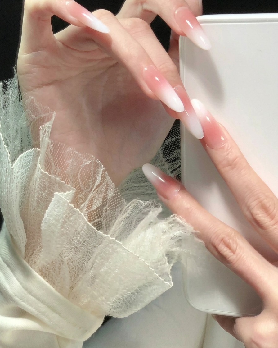 A hand with fingers spread out, showcasing a consistent pink-to-white ombre gradient look on all long, almond-shaped nails.