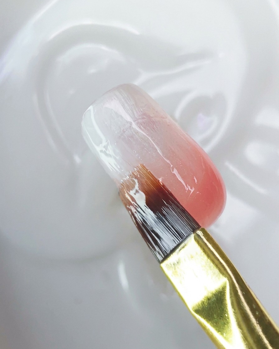 A brush applying a clear top coat over the ombre nail surface.