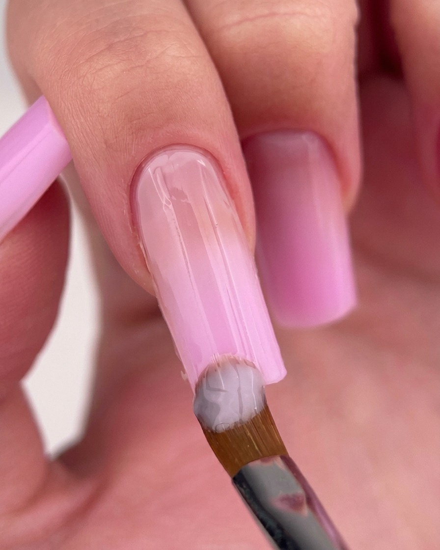 A nail brush applying a light, milky pink gel polish near the cuticle of the nail.