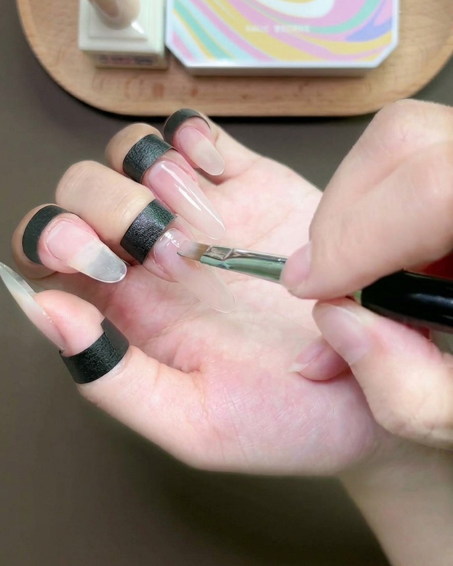 A flat brush applying a thin layer of nude gel onto the middle fingernail.