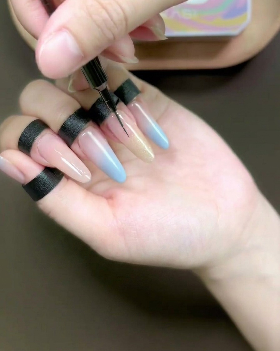 A close-up view of a fine liner brush blending champagne-colored cat-eye gel onto a long, nail-form-extended ring finger.