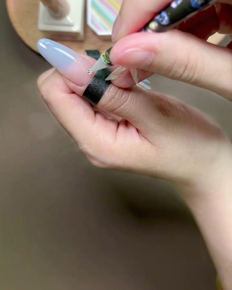A wax pen tool placing a small silver rhinestone onto a bead of glue on a manicured nail.