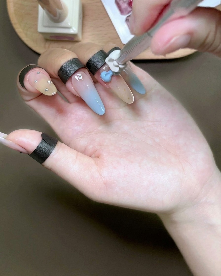 A sponge tool dabbing light blue gel over a 3D white bow charm on a fingernail.