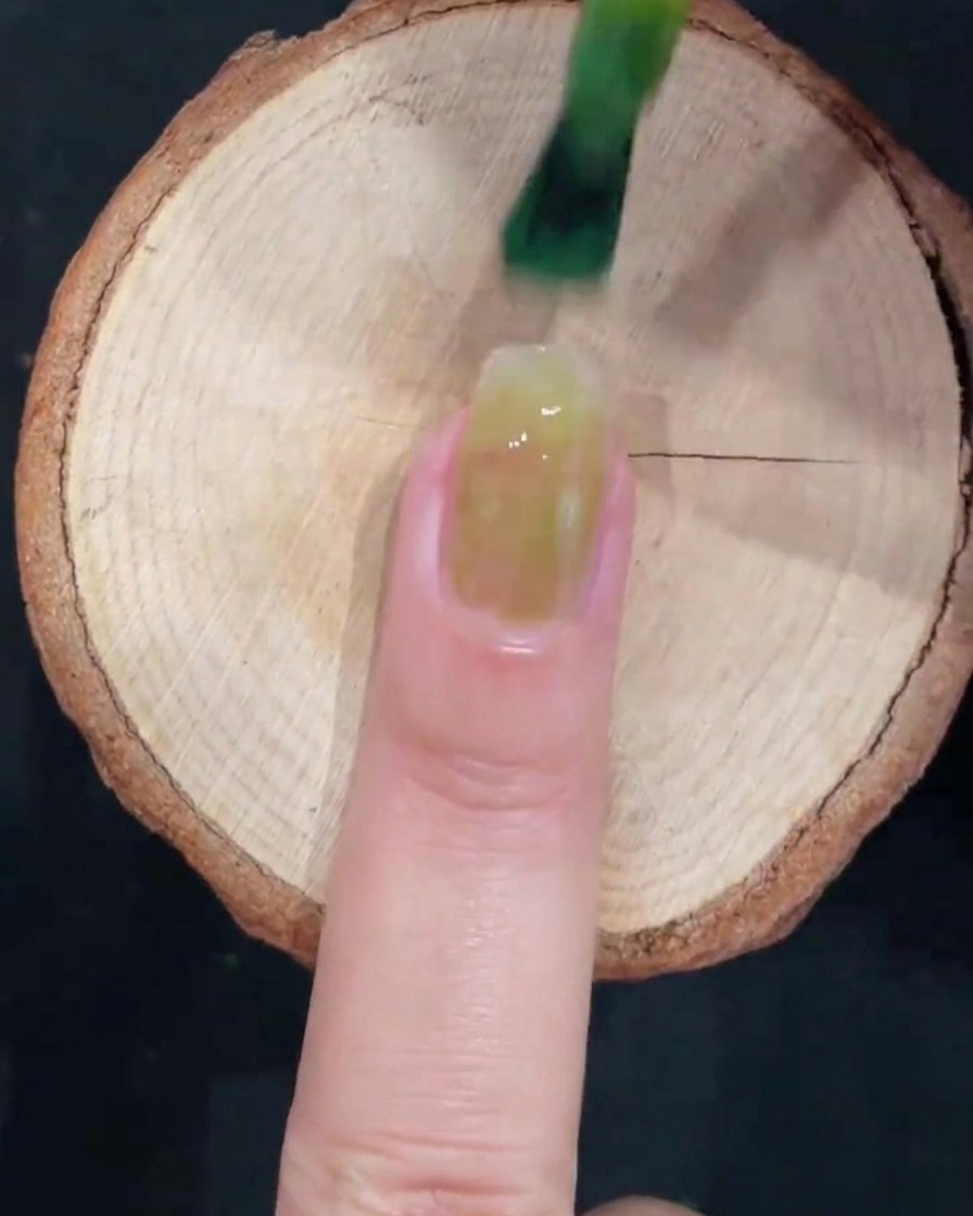 A gel brush applying a sheer icy green polish to the center of a clean natural fingernail over a wooden surface.