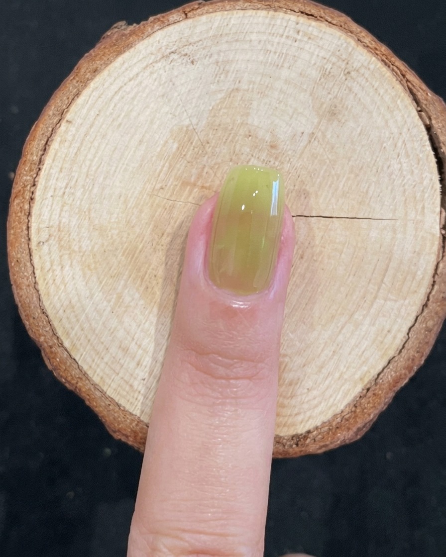 A close-up of a brush applying translucent green gel polish to a natural nail.