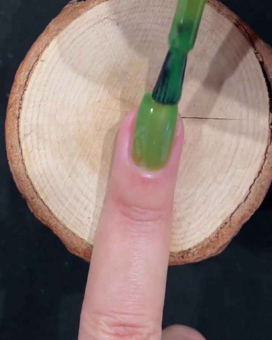 A gel brush completing a full layer of translucent green polish with smooth strokes along the length of the nail.