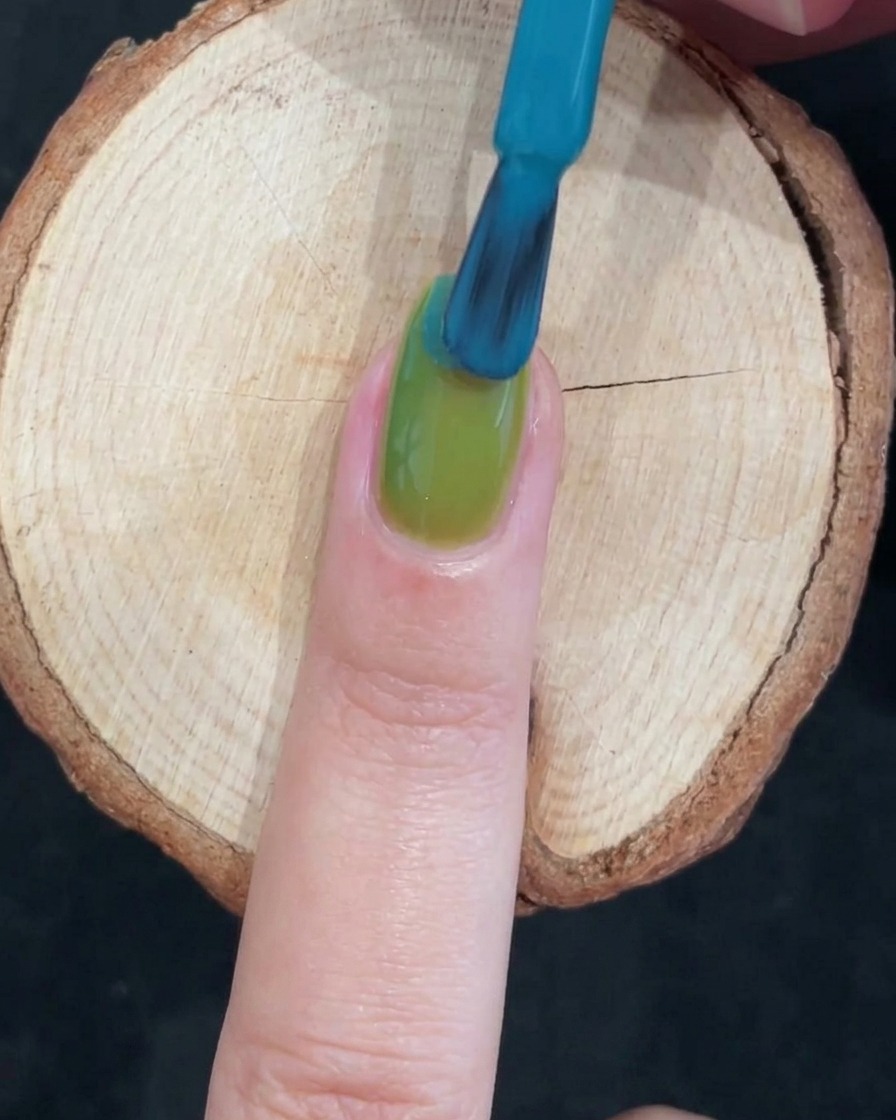 A nail brush applying a vivid blue gel polish over the top half of a glossy green painted nail to create a two-tone gradient.