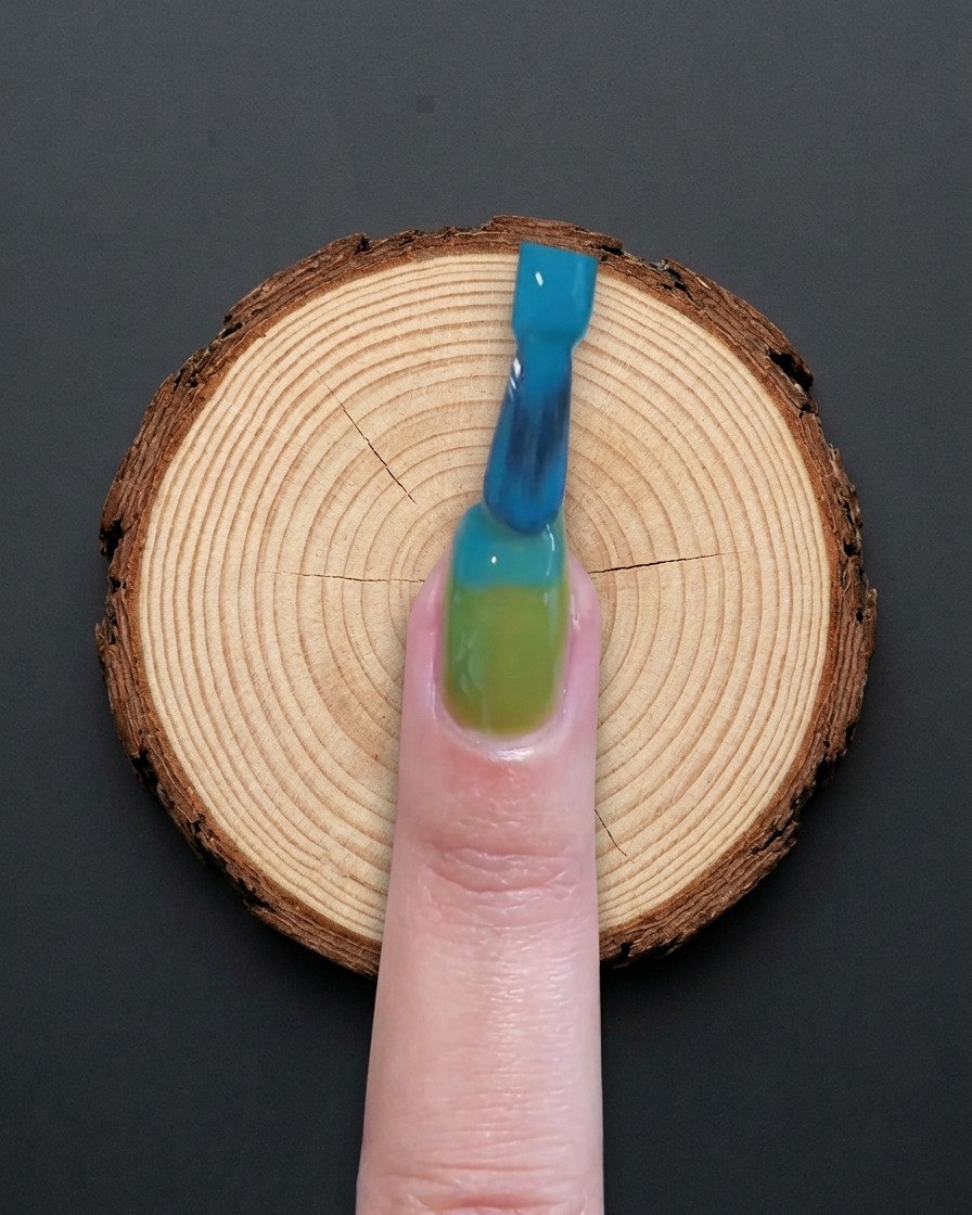 A brush applying blue gel polish to the top half of a green-based nail.