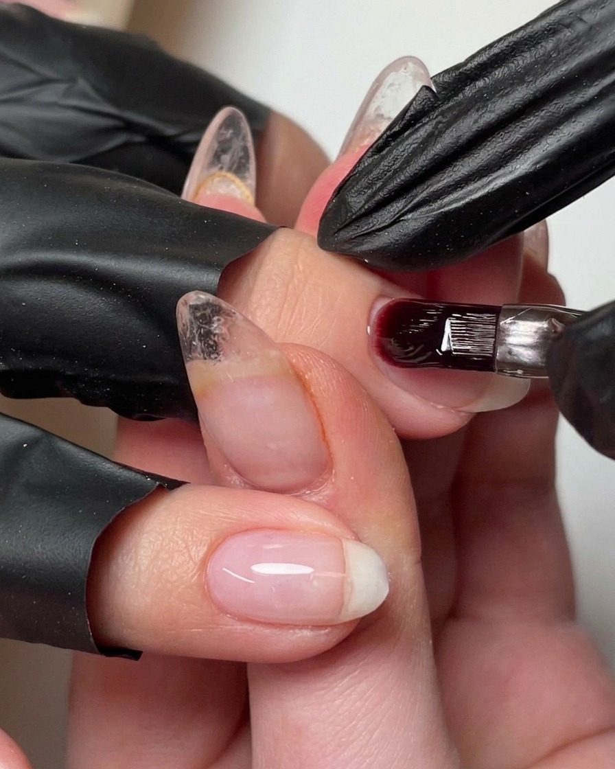 A brush adding a small bead of builder gel to the nail to refine the apex.