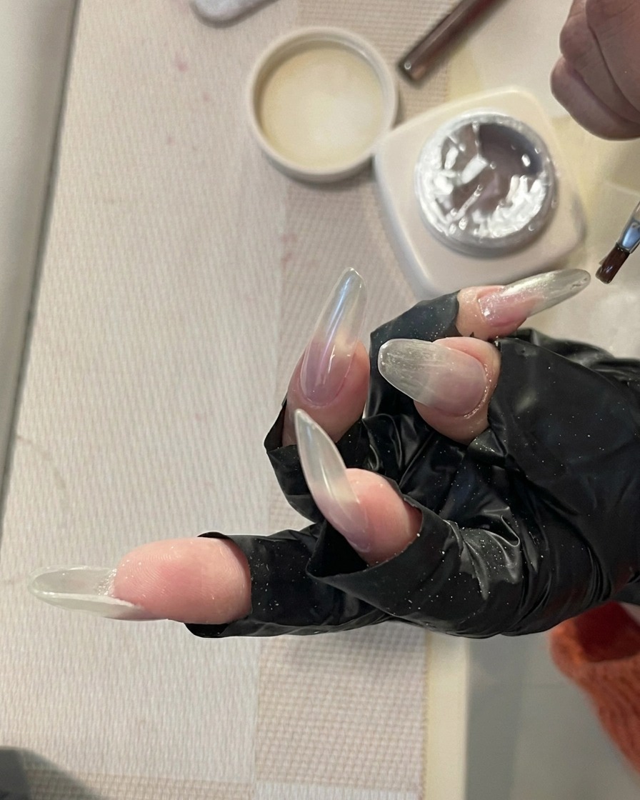 A hand with long, clear nails resting inside a professional UV/LED nail lamp, with the nails illuminated by the curing light.