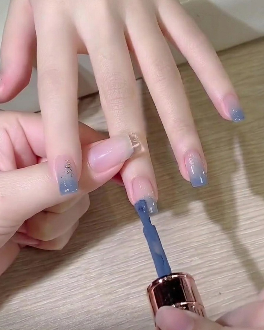 A gel polish bottle brush applying dusty blue color to the tip of a prepped fingernail.