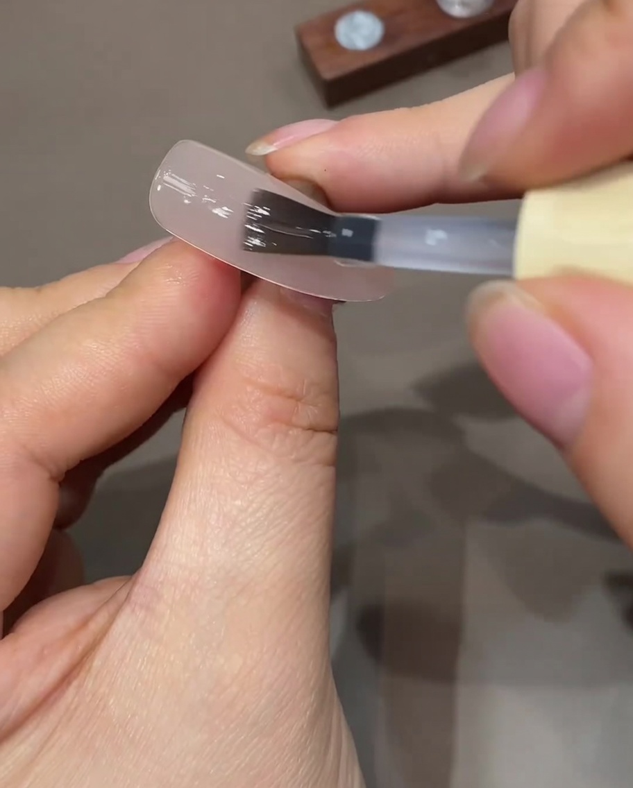 A flat gel brush applying a clear, thin layer of top coat over a sheer nude prepped nail.