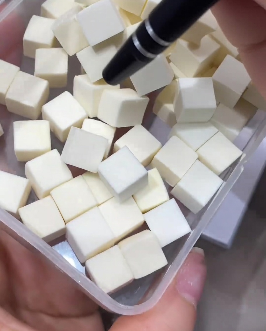 A precision tool picking up a small, square, white makeup sponge cube from a clear plastic container filled with identical clean sponge cubes.