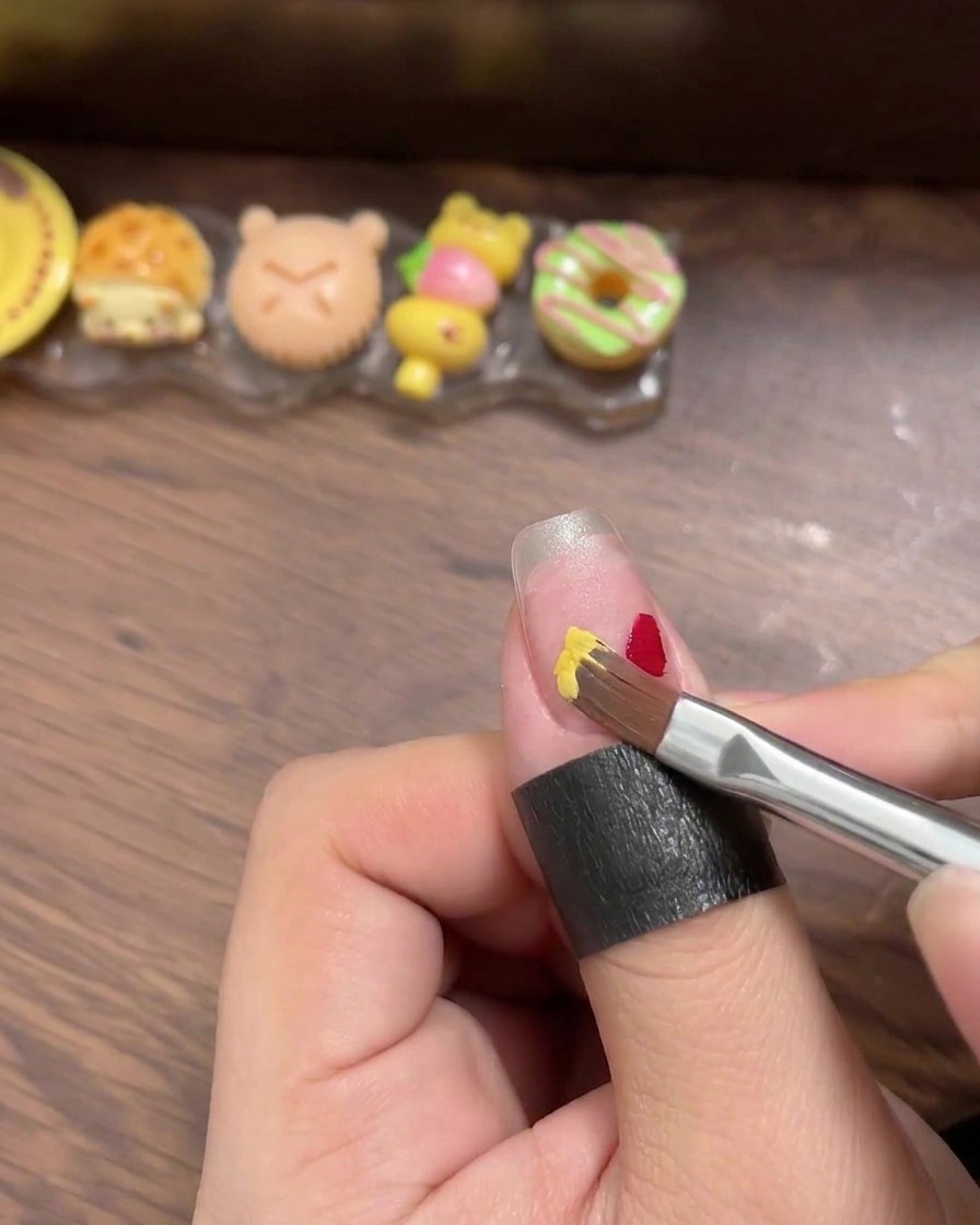 A close-up view of a liner brush applying small, distinct squares of red, yellow, blue, and mint gel polish onto a clear nail tip.