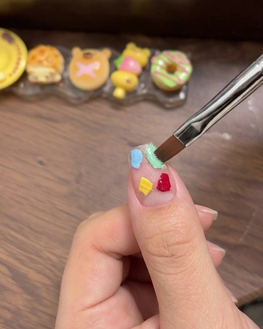 A white cosmetic sponge being used to gently pat and blend four colorful gel squares into a smooth ombre transition on the nail.