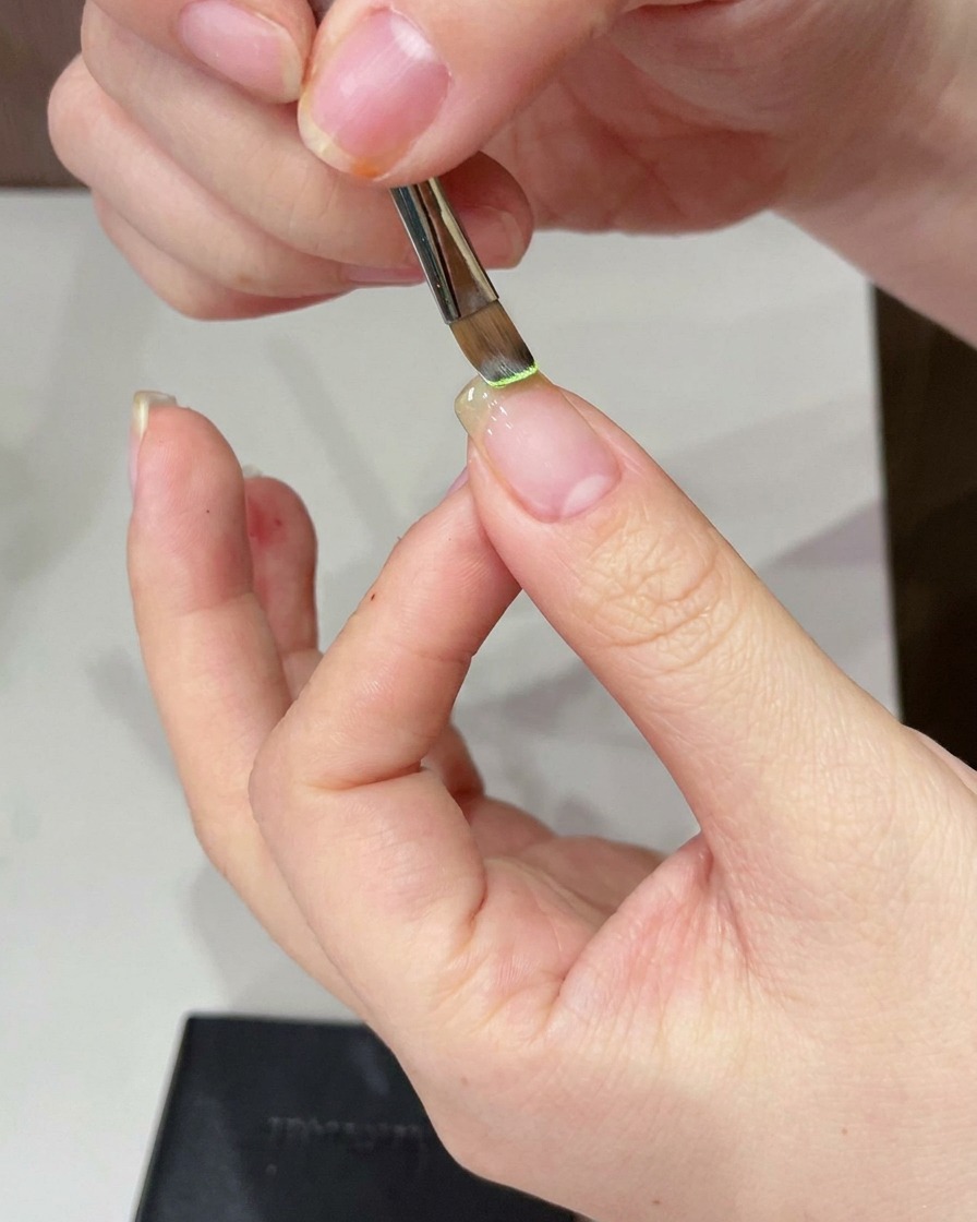 A close-up of a detail brush applying light green gel to a bare nail tip.