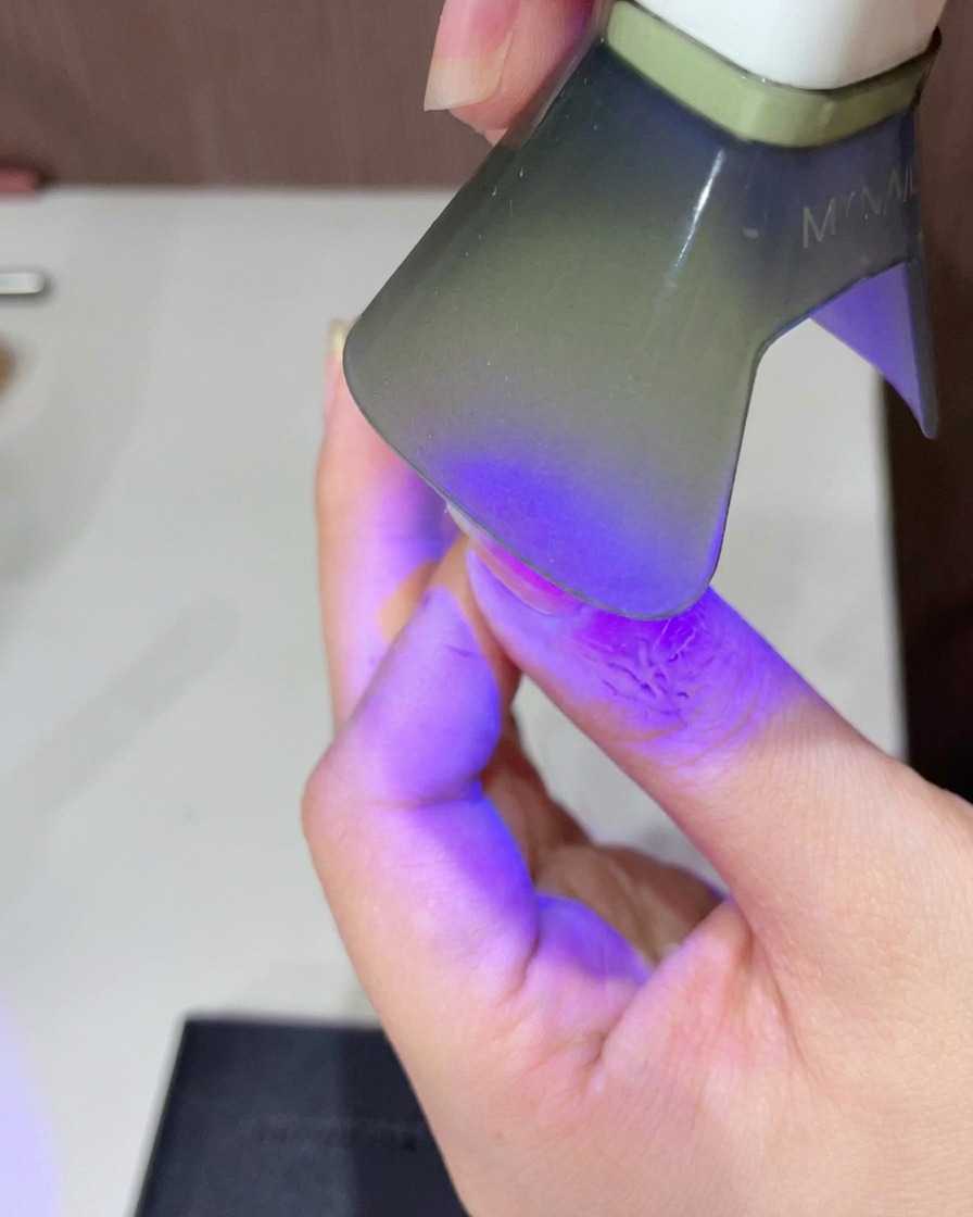 A close-up of a persons finger positioned under a small handheld UV LED lamp to cure the glossy pink gel top coat.
