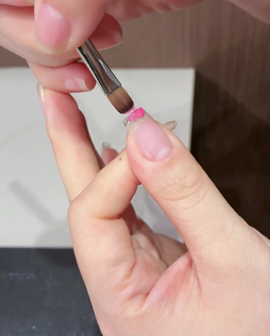 A close-up view of a flat brush applying magenta gel polish specifically to the free edge of a natural nail.