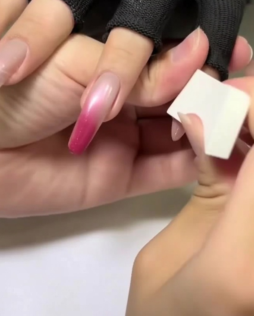 A hand holding a white makeup sponge applying a light rose pink color to the upper part of an almond-shaped nail.