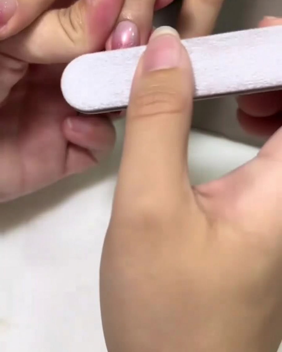 A hand holding a nail file gently buffing the center of a pink polished nail.