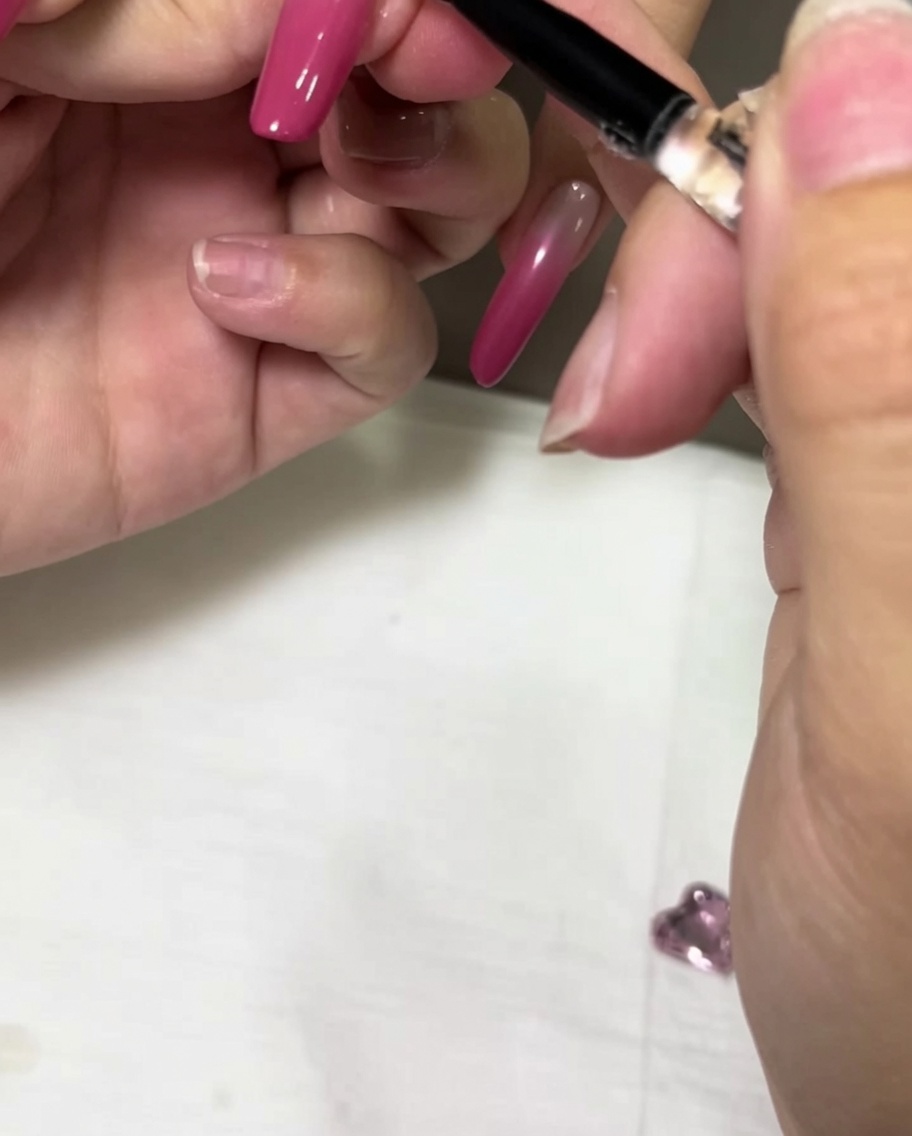 A detail brush applying a bead of clear gel adhesive onto a pink ombre nail.